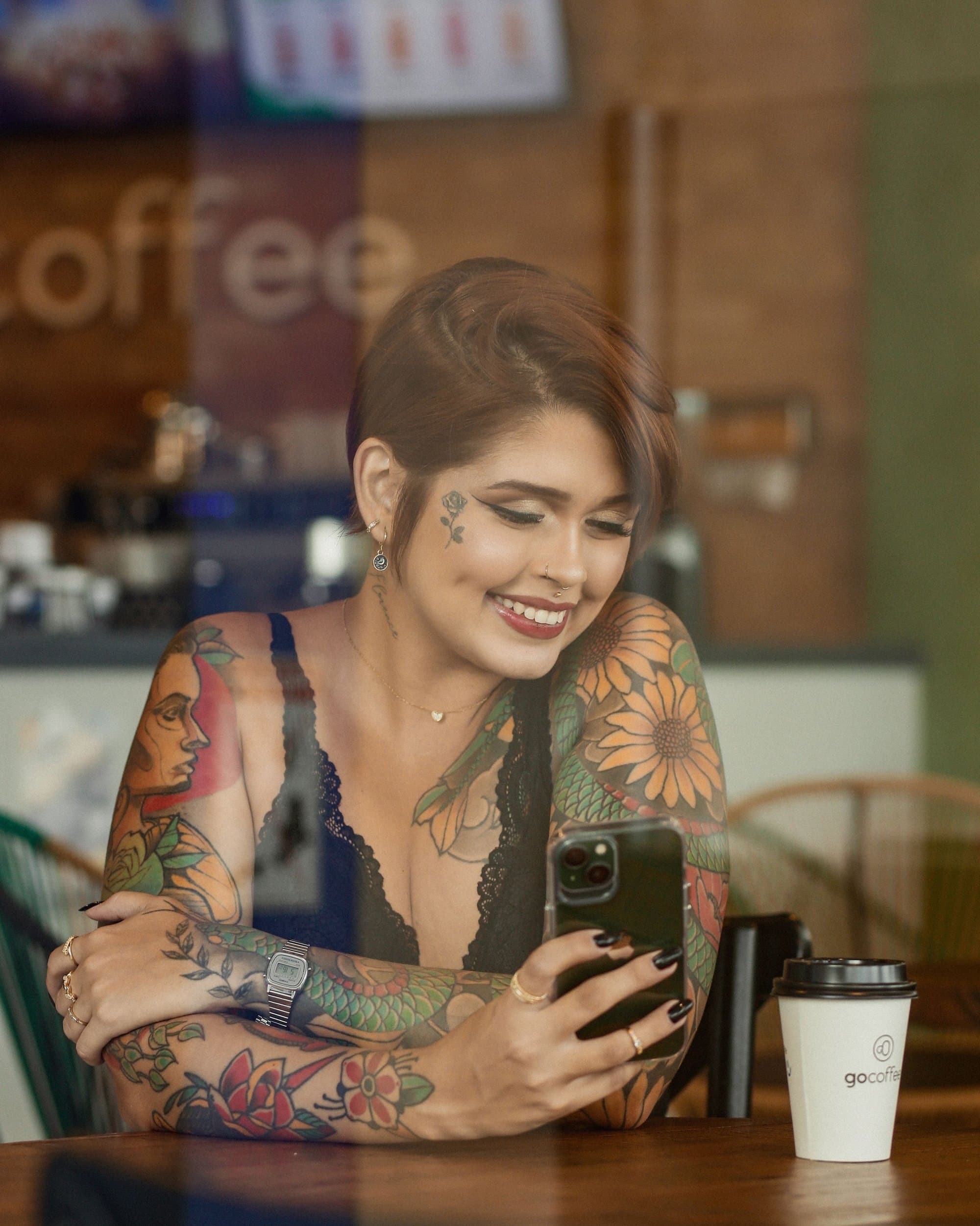 Person mit vielen Tattoos sitzt lächelnd mit dem Handy im Café