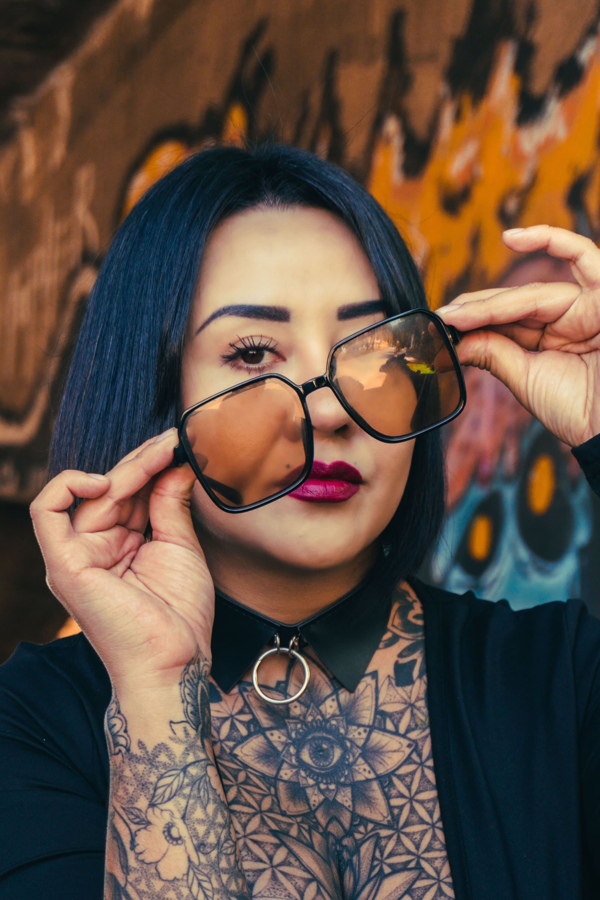 Person mit großer Brille und Tattoos auf der Brust vor einem Graffiti